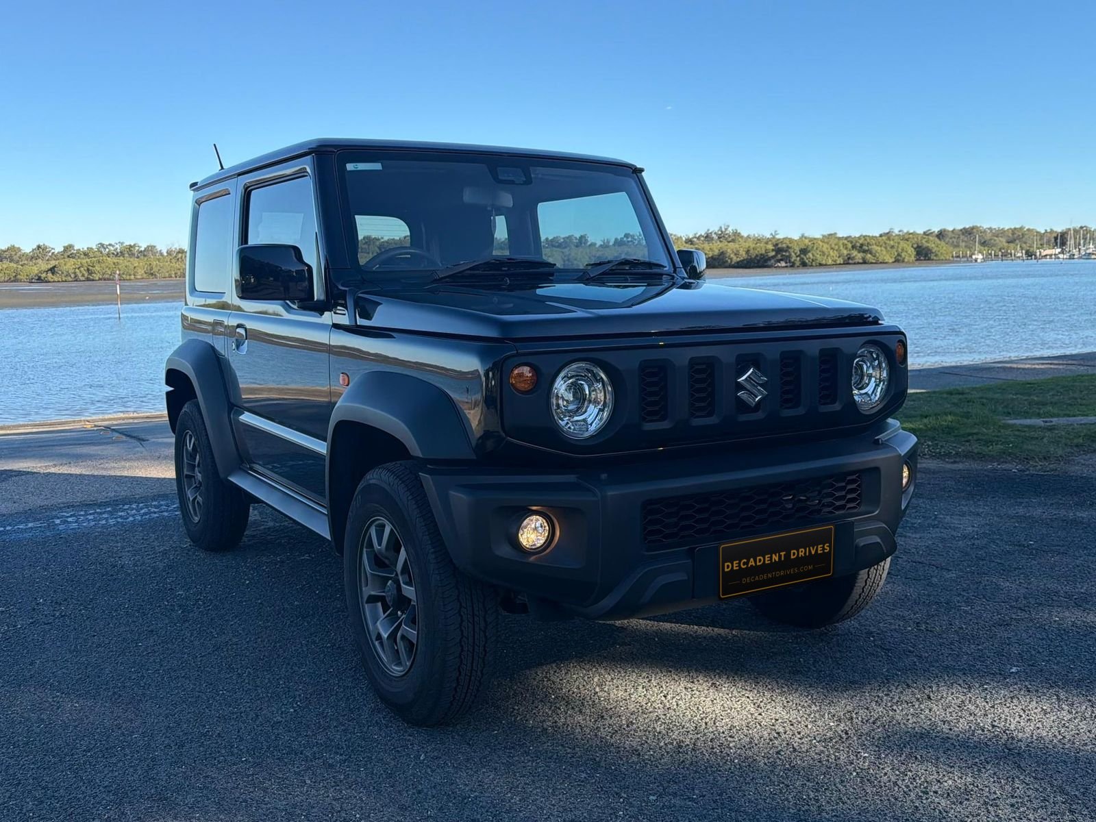Black 2024 Suzuki Jimny small 4WD, side three-quarter view by the water – Decadent Drives hire car, Brisbane